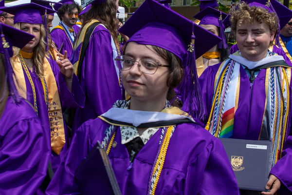 A college graduate walking in a purple commencement robe and mortarboard, surrounded by other college graduates. 