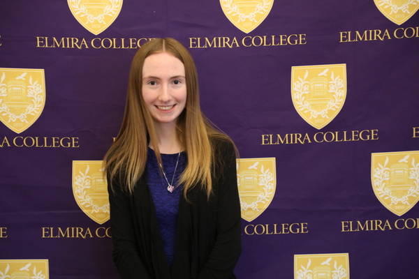 A young woman with long brown hair, wearing a blue dress and black sweater, stands in front of a purple backdrop featuring the Elmira College logo. 