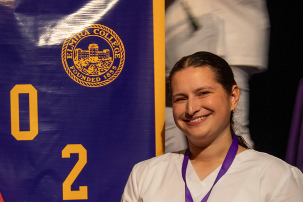 A student in white scrubs stands in front of a purple banner with the Elmira College logo. 