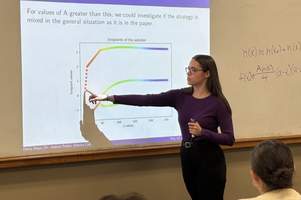 A young woman in a purple top and black pants points to a graph projected on a white board that also has a mathematical formula written on it.  
