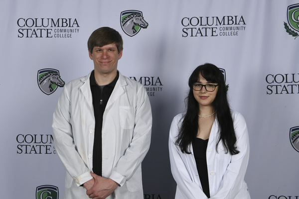 Two people in lab coats stand in front of a white background.
