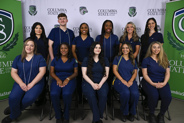 A group of people in dark blue scrubs sits and stands for a photo in front of a white and green background.