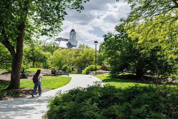 A female student with a backpack walks on an empty tree-lined sidewalk northeast of Love Library on a cloudy day.