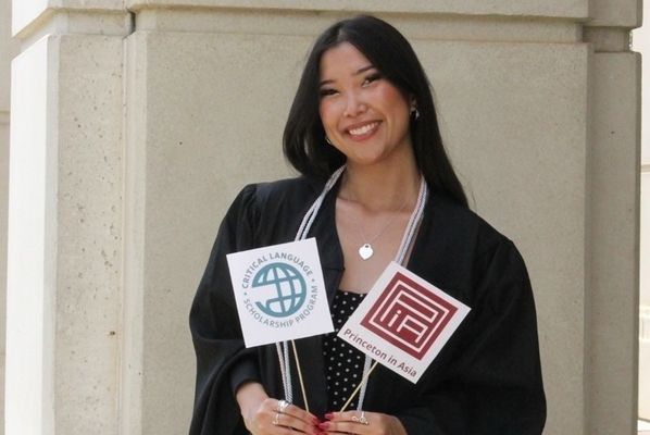 Student stand in front of building in commencement gown with flags for the two programs she accepted scholarships for. 