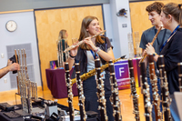 A college student talks to others while holding up a flute, with a display of more musical instruments near her.