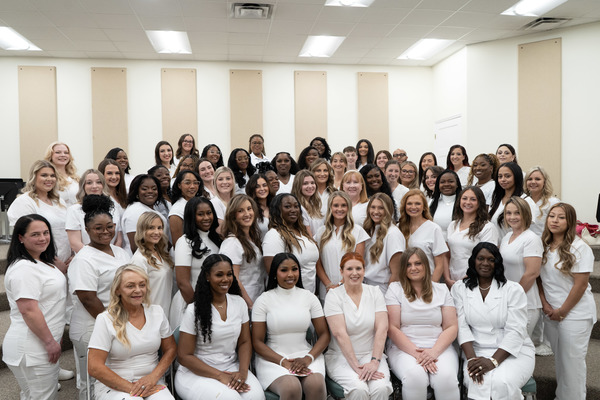 A group of nursing students wearing white scrubs are posing for the camera with a tan and white striped wall behind them.