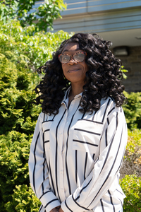 A smiling woman in a striped shirt stands in front of greenery