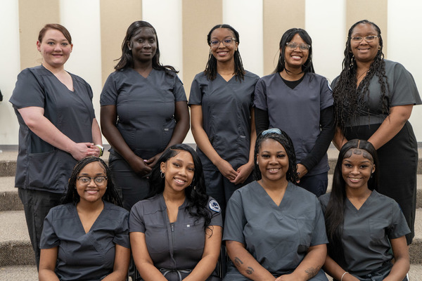 Nine women pose in gray scrubs in front of a tan and white striped wall.