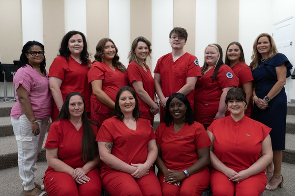 A group of young men and women wearing red scrubs pose in front of a white and tan striped wall.