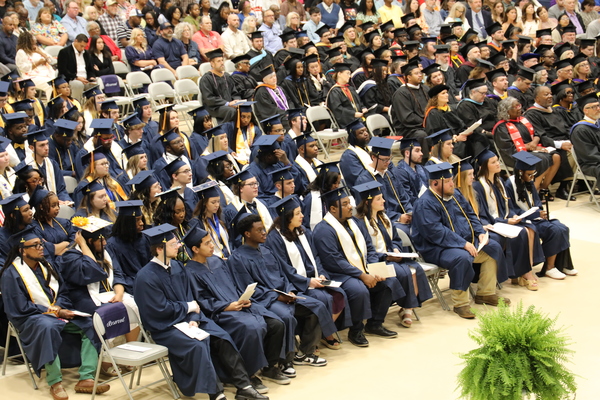 A group of graduates in navy gowns and caps sit listening to a commencement service.