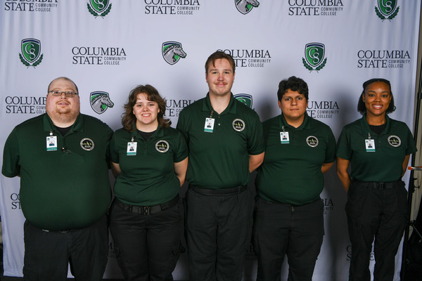 A group of people wearing dark green collar shirts stands in front of a white background.