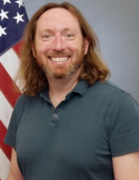Man posing in front of a US flag in civilian clothing.