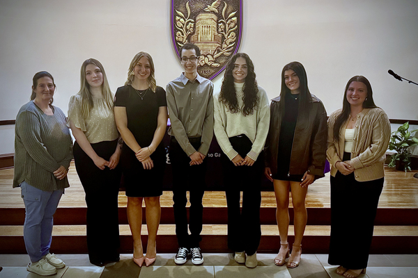 Six students and one professor stand shoulder to shoulder looking at the camera. Behind them is a large Elmira College seal hanging on the wall. 