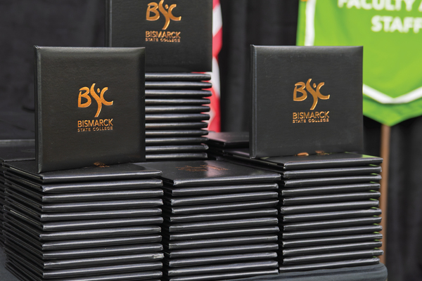 Stacks of black BSC diploma covers with the BSC college logo embossed in gold. Partially visible in the background are a U.S. flag and campus banners.
