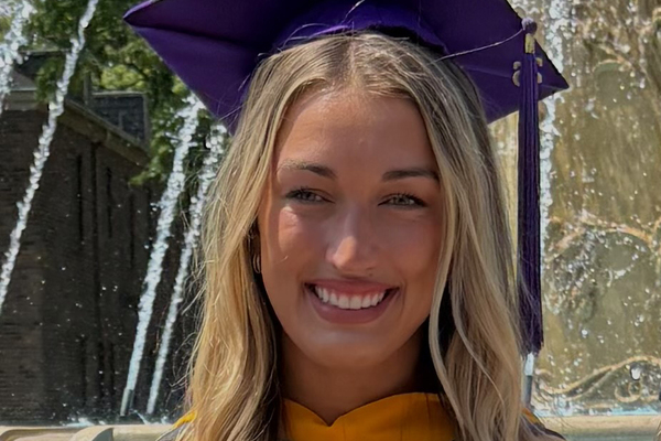 The headshot of a young woman with a big smile wearing a purple mortarboard in front of a fountain. 
