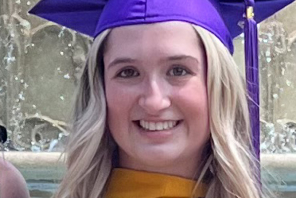 Headshot of a young woman wearing a purple mortarboard standing in front of a fountain. 