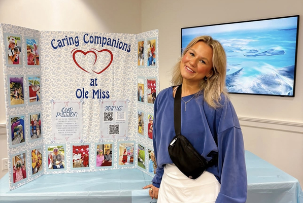 Image of Amber Cecil standing next to a poster that says 'Caring Companions at Ole Miss.'