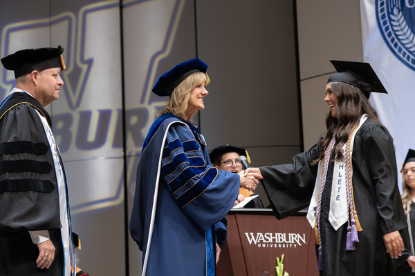 Washburn President Dr. JuliAnn Mazachek shakes hands with a graduate wearing honor cords and a student-athlete stole at commencement.