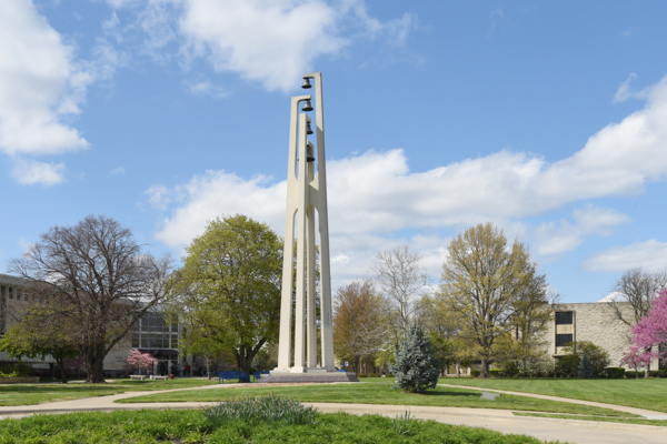 Washburn University's bell tower rises above the campus lawn, surrounded by trees and walkways under a blue sky.