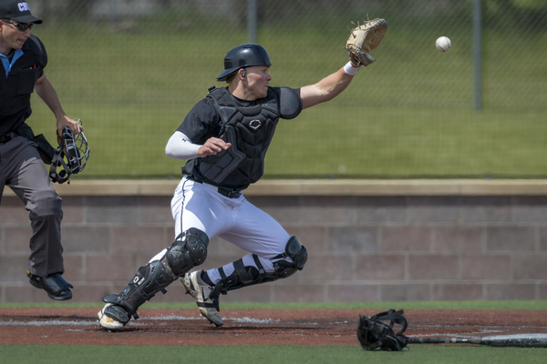 A college baseball catcher catching a baseball with an umpire to their left.