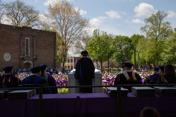 Forefront: silhouettes of Elmira College representatives in their graduate caps and gowns on a stage. Background: a seated audience in purple gowns. 