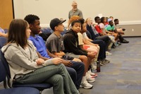 A diverse group of young students sitting in a front row are eagerly listen to a discussion.
