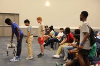 A diverse group of young male students are lined up. One boy dressed in a blue shirt lifts a roboto dog off the ground.