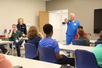 A group of diverse young students sit in a classroom listening while faculty points to someone with a raised hand.