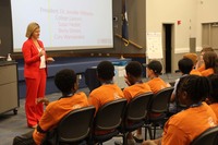 Students all wearing matching orange shirts listen to a woman in a red business suit as she talks. A slideshow is on a screen behind her.