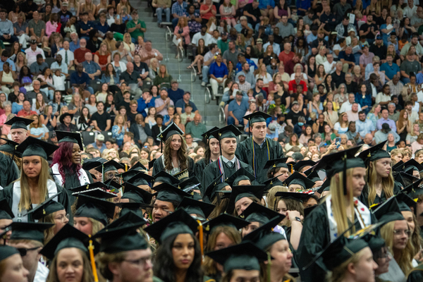 Wide shot of graduates sitting in commencement ceremony, a few standing to be recognized. Spectators sit in the bleachers in the background.