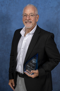 Robert Barry holds his SUNY Canton Hall of Fame Trophy in front of a blue backdrop. 