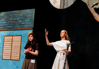 A woman playing the Fairy Godmother holds up her arms while holding a specter. The Cinderella character is next to her.