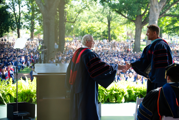 Two men in graduation regalia shake hands on stage in front of a crowd of graduates sitting among a grove of trees.