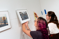 Two students work together to hang framed photography on a gallery wall.