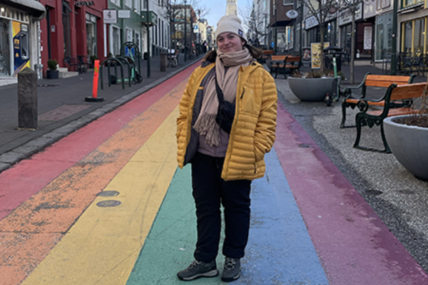 female wearing a yellow puffy coat, cream hat, and scarf standing on a rainbow-painted street