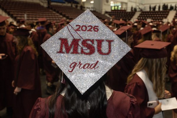 A graduate wearing a sparkling cap with "2026 MSU grad" in red lettering stands among other students in maroon gowns at a large indoor venue.