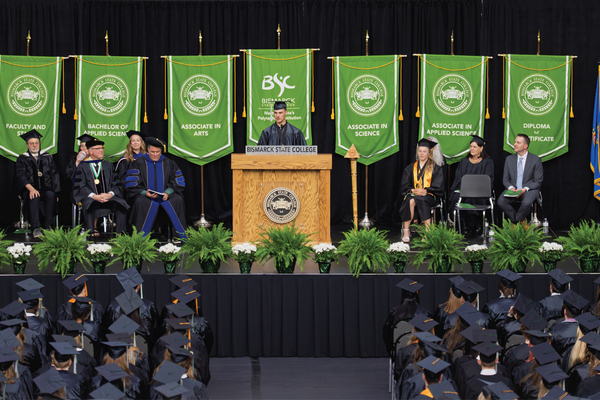 BSC graduate delivers commencement speech at podium as graduates, faculty and green degree banners fill the stage of the Bavendick Center.