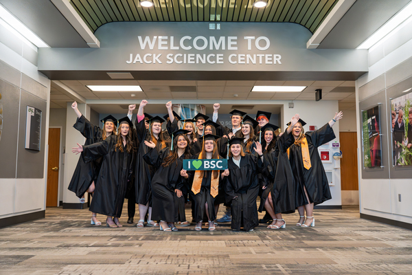 Graduates in caps and gowns celebrate beneath the "Welcome to Jack Science Center" sign, holding an "I ❤️ BSC" sign on campus.