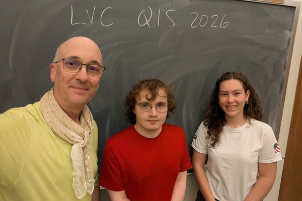 Two students stand with a professor in front of a chalkboard.