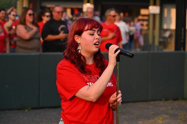 Student sings at a microphone as crowd members have their hands over their hearts. 