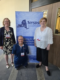 Nichelle Burnett, John Robinson and Tamara Mariotti pose in front of a NYSDSC banner during a conference.