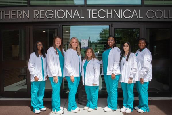 Seven Surgical Technology graduates in teal scrubs and white lab coats stand together outside the entrance to Southern Regional Technical College.