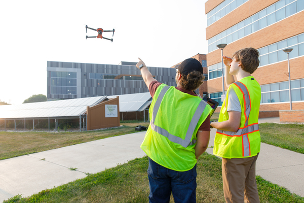 Two students in safety vests operate a drone outside on campus, gaining hands-on flight experience near buildings and solar panels.