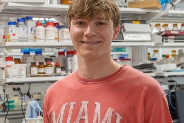 student wearing red t-shirt with the word Miami in white while standing in lab/pharmacy setting