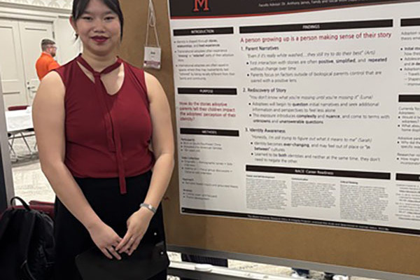 Student wearing a sleeveless burgundy blouse standing in front of a research poster