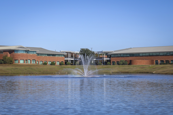 Brick campus building with a pond with a spraying fountain in front, under clear blue sky, surrounded by manicured lawn.