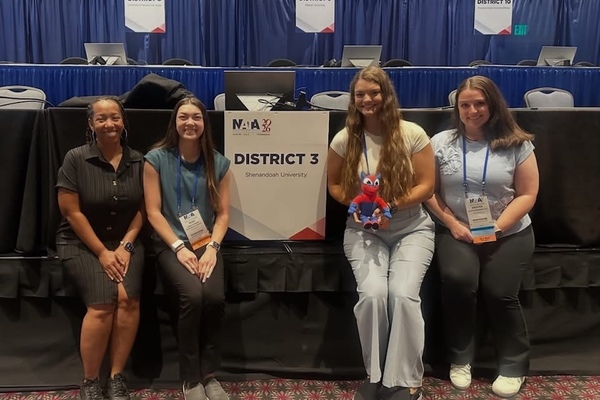 Three Shenandoah University students pose with their program director with a sign that reads "District 3".