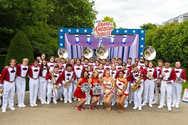 Members of the Marching back stand in front of a stage with a sign that reads Grand American Jubilee.