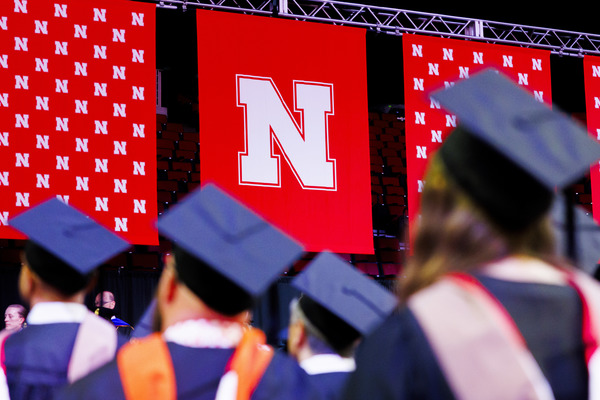 Husker graduates in black robes and mortar boards face the stage at Pinnacle Bank Arena. Nebraska-branded red banners hang behind the stage.