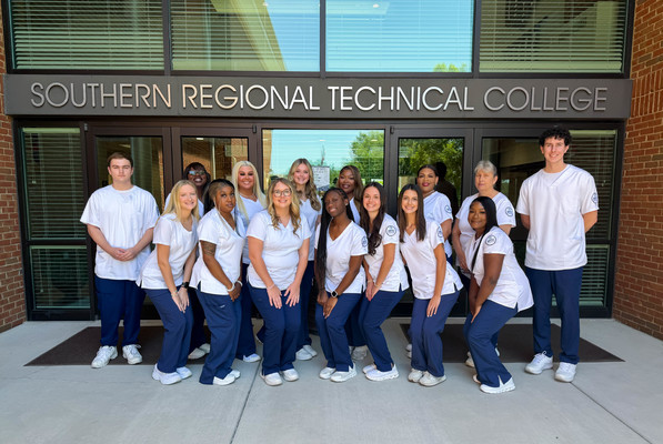 Nursing graduates in blue scrub pants and white scrub tops stand outside the entrance to SRTC
